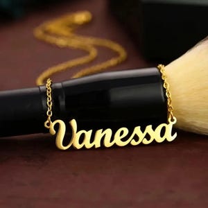May include: A gold-colored necklace with the name "Vanessa" in a cursive font. The necklace is on a gold chain. The background includes a black object and a makeup brush with a beige head.