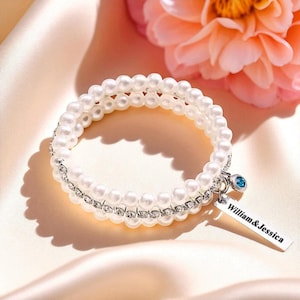 Op de afbeelding: Een dubbelstrengs parelarmband met een zilveren band van kleine, heldere kristallen. Een kleine, rechthoekige bedel met de namen "William & Jessica" hangt aan de armband, samen met een kleine blauwe edelsteen. De armband is geplaatst tegen een zachte, satijnen achtergrond.