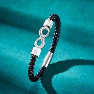 May include: A black braided leather bracelet with a silver infinity symbol and accents. The infinity symbol has the names "Tina & David" engraved on it. The word "LOVE" is engraved on one of the silver accents.