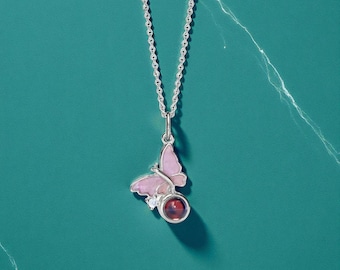 May include: A silver necklace with a butterfly pendant. The butterfly pendant is pink with a red gemstone and a small clear stone. The necklace is displayed on a teal surface.