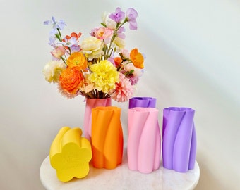 Puede incluir: Una colección de coloridos jarrones en tonos amarillo, naranja, rosa y morado. Los jarrones tienen un diseño retorcido único. Un ramo de flores en un jarrón rosa se encuentra detrás de los otros jarrones. Las flores son en tonos amarillo, naranja, rosa y morado.