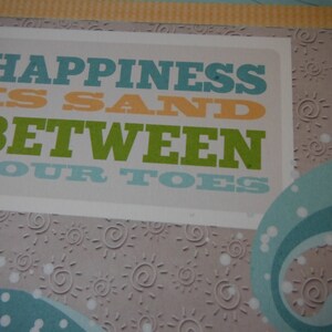 May include: A light gray background with a white rectangle featuring the text "Happiness is sand between your toes" in blue, orange, and green lettering. The background is decorated with white sun shapes.