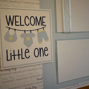 May include: A beige scrapbook page with a blue and white patterned background. The page features a white card with the text "WELCOME little one" in black lettering. The card has a graphic of three baby clothes hanging on a clothesline. The clothes are a bib, a onesie, and pants, all in blue with yellow accents.