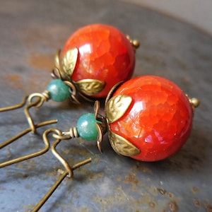 Op de afbeelding: Een paar bungelende oorbellen met grote, ronde, gebarsten oranje kralen bekroond met antiek messing blad-vormige kraalkappen. Elke oorbel bevat een kleine, gladde, ronde groene kraal en hangt aan een antiek messing oorhaakje.