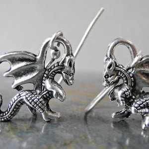 May include: A pair of silver dragon earrings with a detailed design. The dragons have wings, scales, and a curled tail. The earrings are made of metal and have a hook fastening.