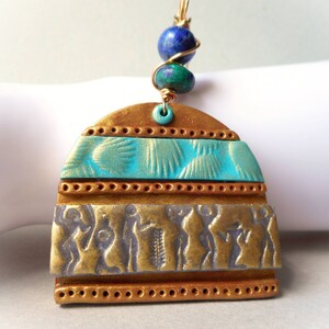 May include: A gold, turquoise, and silver pendant with a geometric design and a beaded top. The pendant features a repeating pattern of small circles and a band of figures.