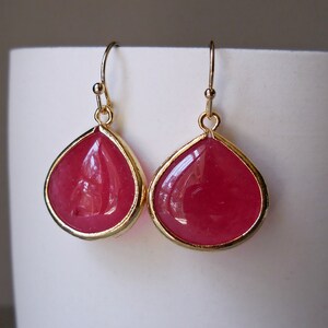 May include: A pair of teardrop-shaped earrings with a vibrant pink gemstone center. The gemstones are encased in a gold-colored metal frame and suspended from gold-colored ear wires. The earrings are displayed against a white background.