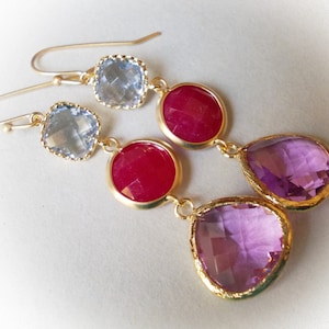 May include: Gold dangle earrings with three gemstone drops. The top drop is a light blue square, the middle drop is a red circle, and the bottom drop is a purple teardrop.