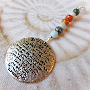 Könnte beinhalten: Silberfarbener Anhänger mit gehämmertem Muster und hebräischen Schriftzeichen. Der runde Anhänger hat eine Kette aus bunten Perlen in Blau-, Orange- und Grüntönen. Oben befindet sich eine kleine Silberschlaufe.