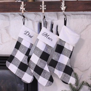 Puede incluir: Tres calcetines de Navidad de cuadros negros y blancos con ribete de piel blanca. Los calcetines están colgados de una repisa de madera con ganchos plateados. Los calcetines están personalizados con los nombres "Papá", "Mamá" y un tercer calcetín sin nombre.
