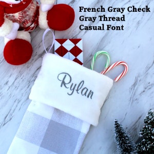 May include: White Christmas stocking with a gray and white buffalo plaid pattern. The stocking has a white cuff with the name "Rylan" embroidered in gray thread. The stocking is filled with candy canes and a red and white gift box.