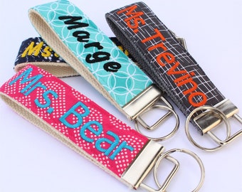 May include: Four personalized keychains with different fabric patterns and embroidered names. The keychains are teal, pink, gray, and black with white and orange embroidery.