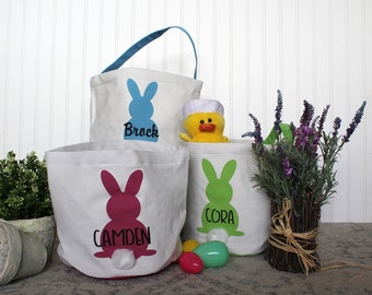 Puede incluir: Tres cestas de Pascua de lona blanca con siluetas de conejos y nombres personalizados: Brock, Camden y Cora. Las cestas están llenas de huevos de Pascua y un pollito de peluche amarillo.