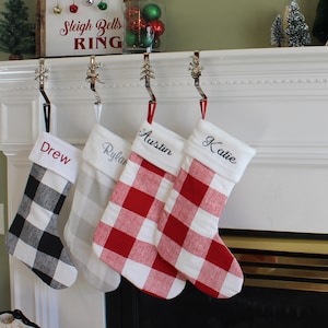 May include: Four personalised Christmas stockings hanging on a white mantelpiece. The stockings are red and white buffalo plaid, grey and white buffalo plaid, and black and white buffalo plaid. The stockings are embroidered with the names "Drew", "Rylan", "Austin", and "Katie".