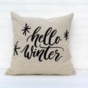 May include: A beige throw pillow with black embroidered text that reads "*hello* winter*"