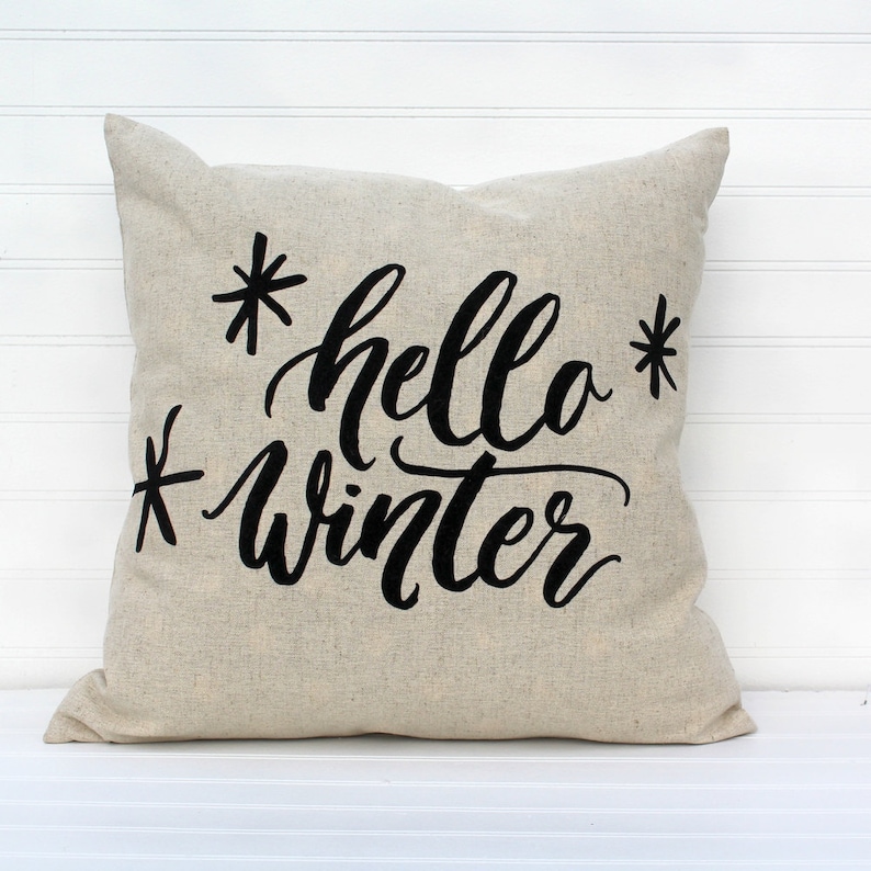 May include: A beige throw pillow with black embroidered text that reads "*hello* winter*"