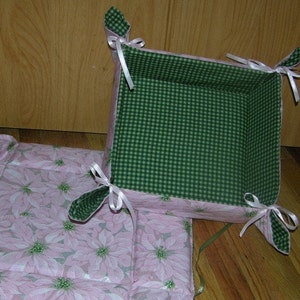 May include: A green and white gingham fabric basket with white ribbon handles. The basket is sitting on a pink fabric with a floral pattern.