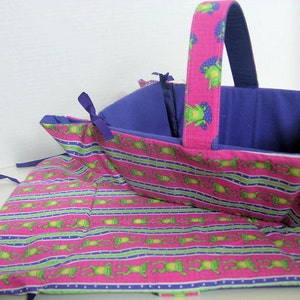 May include: A pink and purple fabric basket with a frog print and a purple lining. The basket has a handle and is folded open to show the interior.