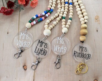 May include: Four clear acrylic name tags with black text on a wooden background. The name tags are attached to beaded lanyards with a variety of colors. The name tags read "Mrs. Valdez", "Mrs. Lytle", "Ms. White", and "Mrs. Rutledge".