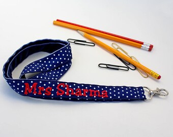 Puede incluir: Un cordón azul y blanco con lunares con un nombre bordado en rojo "Mrs. Sharma" y un clip giratorio plateado.