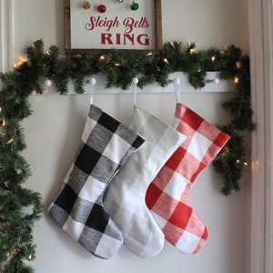 May include: Three Christmas stockings hanging on a white wall. The stockings are made of fabric and have a buffalo plaid pattern. The stockings are black and white, gray and white, and red and white.