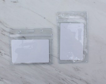 May include: Two clear plastic badge holders with white card inserts. The holders have a slot at the top for a lanyard.