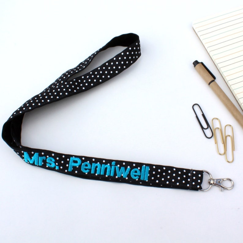 Puede incluir: Lanyard de lunares blanco y negro con un nombre bordado en azul "Mrs. Penniwell" y un clip giratorio plateado.
