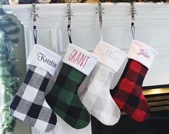 May include: Four personalized Christmas stockings with buffalo plaid patterns in black and white, green and white, gray and white, and red and black. The stockings are hung on a white fireplace mantel with silver hooks. The names "Kristin", "Grant", "Mom", and "Jason" are embroidered on the stockings.