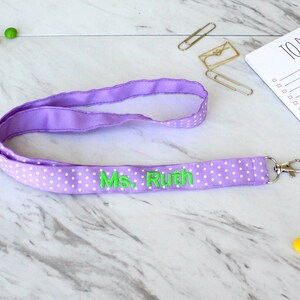 Puede incluir: Lanyard morado con lunares blancos y el texto "Ms. Ruth" bordado en verde.