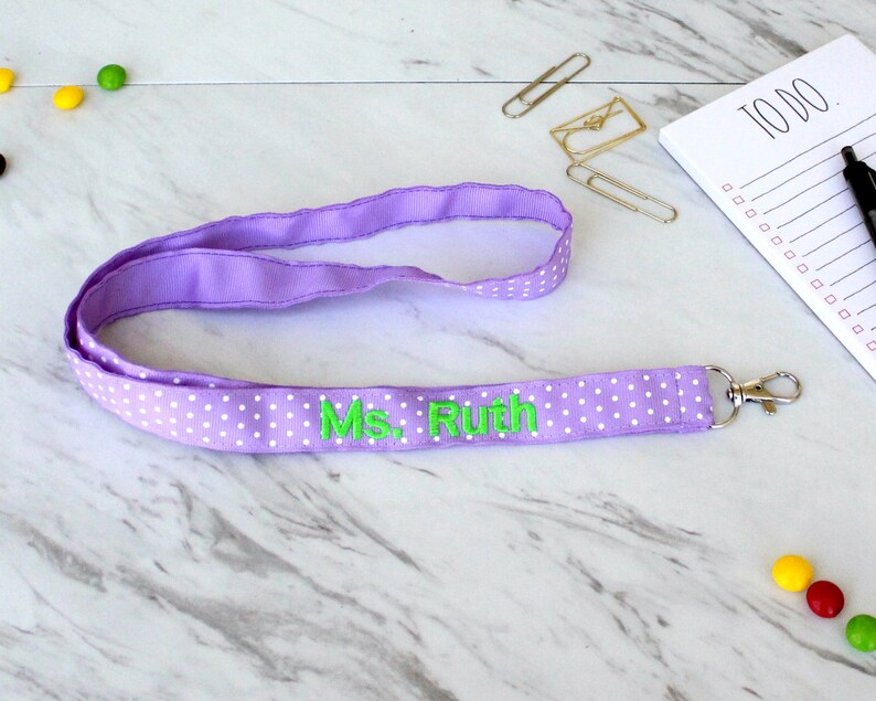 Puede incluir: Lanyard morado con lunares blancos y el texto "Ms. Ruth" bordado en verde.