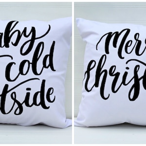 May include: Two white throw pillows with black text. The first pillow says "Baby it's cold outside". The second pillow says "Merry Christmas".