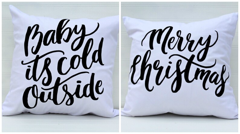 May include: Two white throw pillows with black text. The first pillow says "Baby it's cold outside". The second pillow says "Merry Christmas".