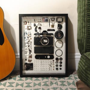 Puede incluir: Una exhibición enmarcada de una cámara Yashica FX-3 desmontada. El cuerpo y la lente negros de la cámara están rodeados de varias piezas pequeñas, tornillos y componentes, todos dispuestos sobre un fondo blanco. El marco es negro.