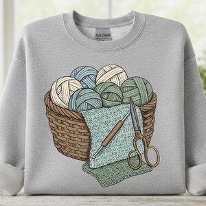Puede incluir: Sudadera gris claro con un gráfico de una cesta de mimbre llena de ovillos de lana en tonos azules, verdes y crema. También se representan un ganchillo, unas tijeras y un cuadrado de ganchillo. La sudadera tiene mangas enrolladas.