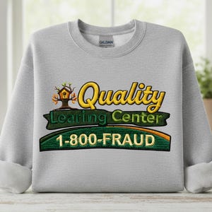 Könnte beinhalten: Ein hellgraues Sweatshirt mit dem gestickten Text "Quality Learning Center" in Gelb und Grün sowie der Telefonnummer "1-800-FRAUD" in Grün und Orange. Das Sweatshirt hat einen Rundhalsausschnitt und hochgekrempelte Ärmel.