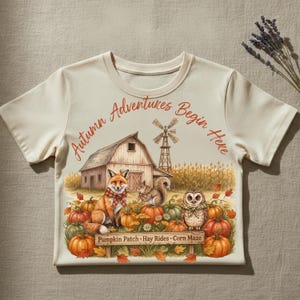 Könnte beinhalten: Ein cremefarbenes T-Shirt mit einer Grafik aus Scheune, Windmühle und Maisfeld. Ein Fuchs, ein Eichhörnchen und eine Eule sitzen zwischen Kürbissen. Der Text lautet: Autumn Adventures Begin Here, Pumpkin Patch, Hay Rides, Corn Maze in Orange und Braun.
