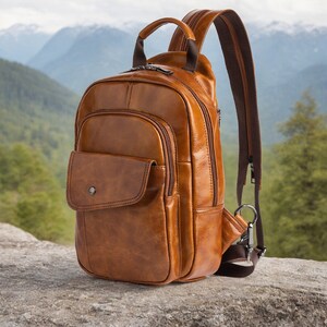 Pode incluir: Uma mochila de couro marrom com uma única alça e um bolso frontal. A mochila está sentada em uma rocha com um fundo verde e marrom.