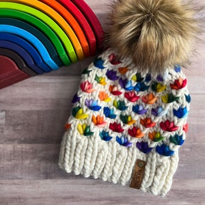 May include: A white knit beanie with a rainbow flower pattern and a brown faux fur pom pom. The beanie has a brown leather tag that says "Made for the World".