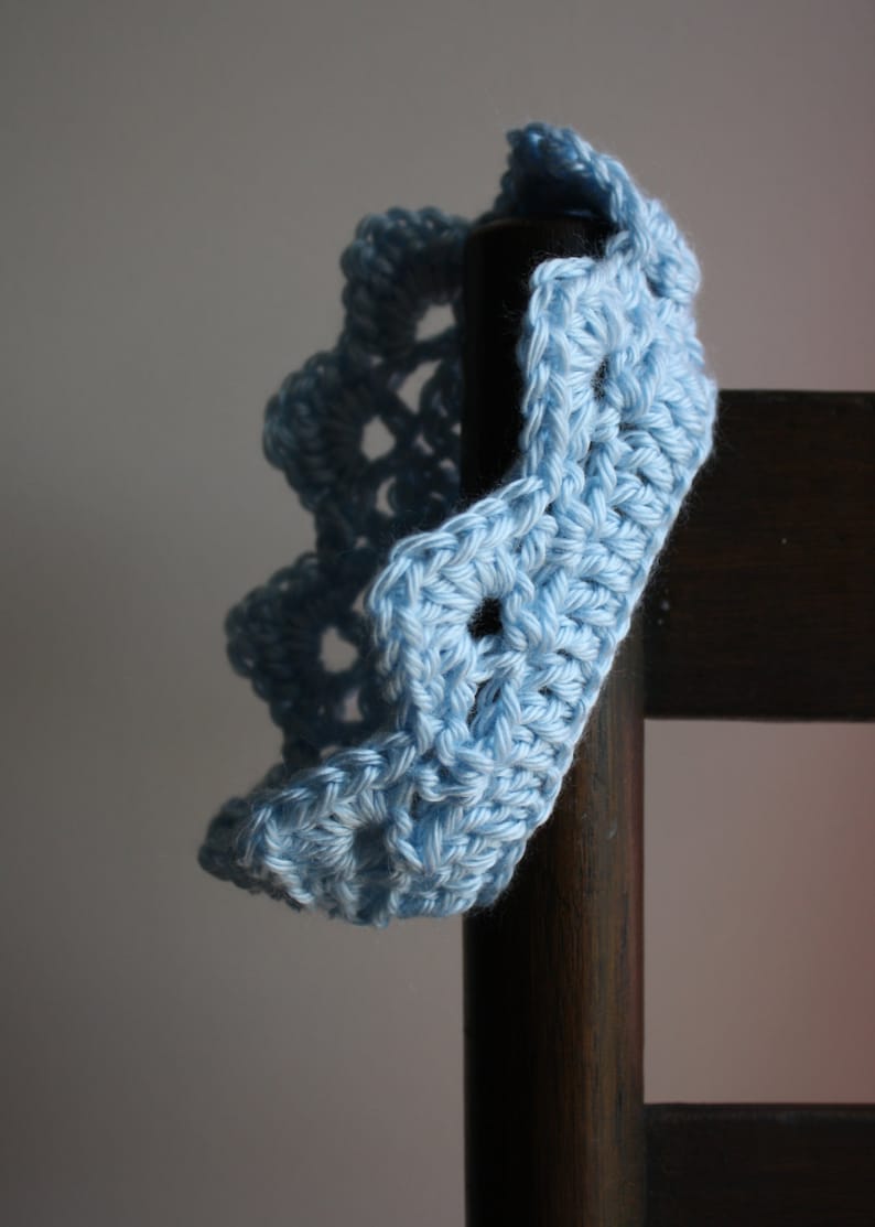May include: A light blue crocheted headband with a scalloped edge. The headband is draped over a wooden chair.