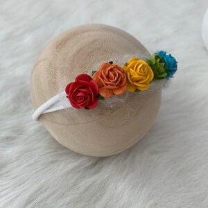K&ouml;nnte beinhalten: Ein wei&szlig;es Stirnband mit einem Regenbogen aus Stoffblumen. Die Blumen sind rot, orange, gelb, gr&uuml;n und blau.