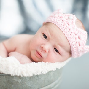 May include: A newborn baby wearing a pink crocheted crown headband lies in a white fuzzy bucket. The baby is looking at the camera with a serious expression.