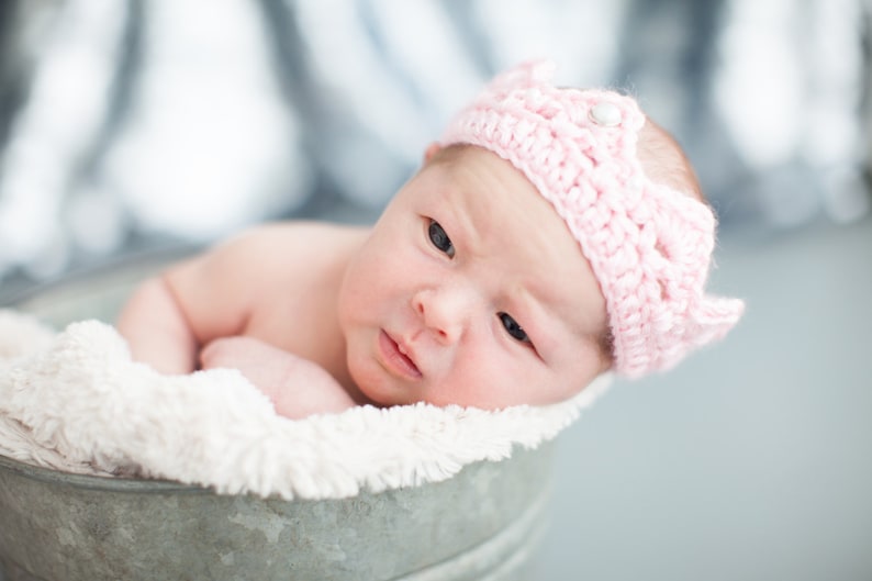 May include: A newborn baby wearing a pink crocheted crown headband lies in a white fuzzy bucket. The baby is looking at the camera with a serious expression.