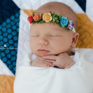 K&ouml;nnte beinhalten: Ein neugeborenes Baby tr&auml;gt ein wei&szlig;es Stirnband mit einem Regenbogen aus Filzblumen. Das Baby schl&auml;ft friedlich auf einer bunten Steppdecke.