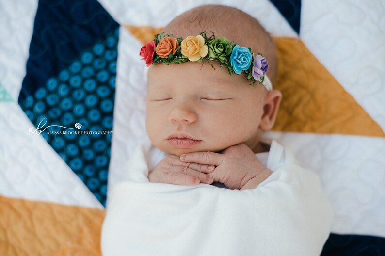 K&ouml;nnte beinhalten: Ein neugeborenes Baby tr&auml;gt ein wei&szlig;es Stirnband mit einem Regenbogen aus Filzblumen. Das Baby schl&auml;ft friedlich auf einer bunten Steppdecke.