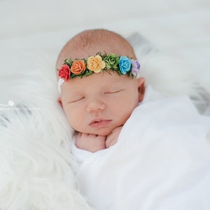 K&ouml;nnte beinhalten: Ein neugeborenes Baby tr&auml;gt ein wei&szlig;es Stirnband mit einem Regenbogen aus bunten Stoffblumen. Das Baby schl&auml;ft friedlich mit den H&auml;nden unter dem Kinn.
