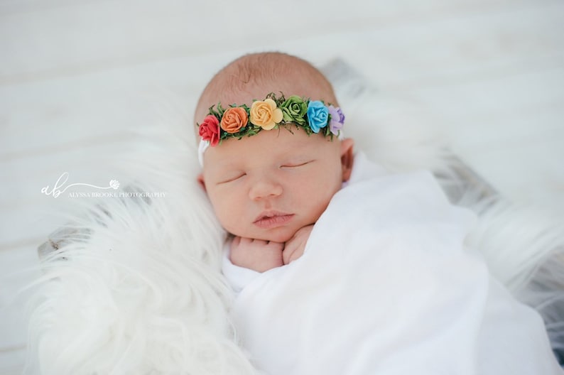 K&ouml;nnte beinhalten: Ein neugeborenes Baby tr&auml;gt ein wei&szlig;es Stirnband mit einem Regenbogen aus bunten Stoffblumen. Das Baby schl&auml;ft friedlich mit den H&auml;nden unter dem Kinn.