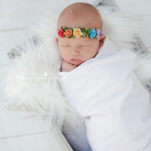K&ouml;nnte beinhalten: Ein neugeborenes Baby mit einem Regenbogenblumen-Stirnband schl&auml;ft in einem wei&szlig;en, mit Fell ausgekleideten Korb. Das Baby ist in eine wei&szlig;e Decke gewickelt und hat die Augen geschlossen.