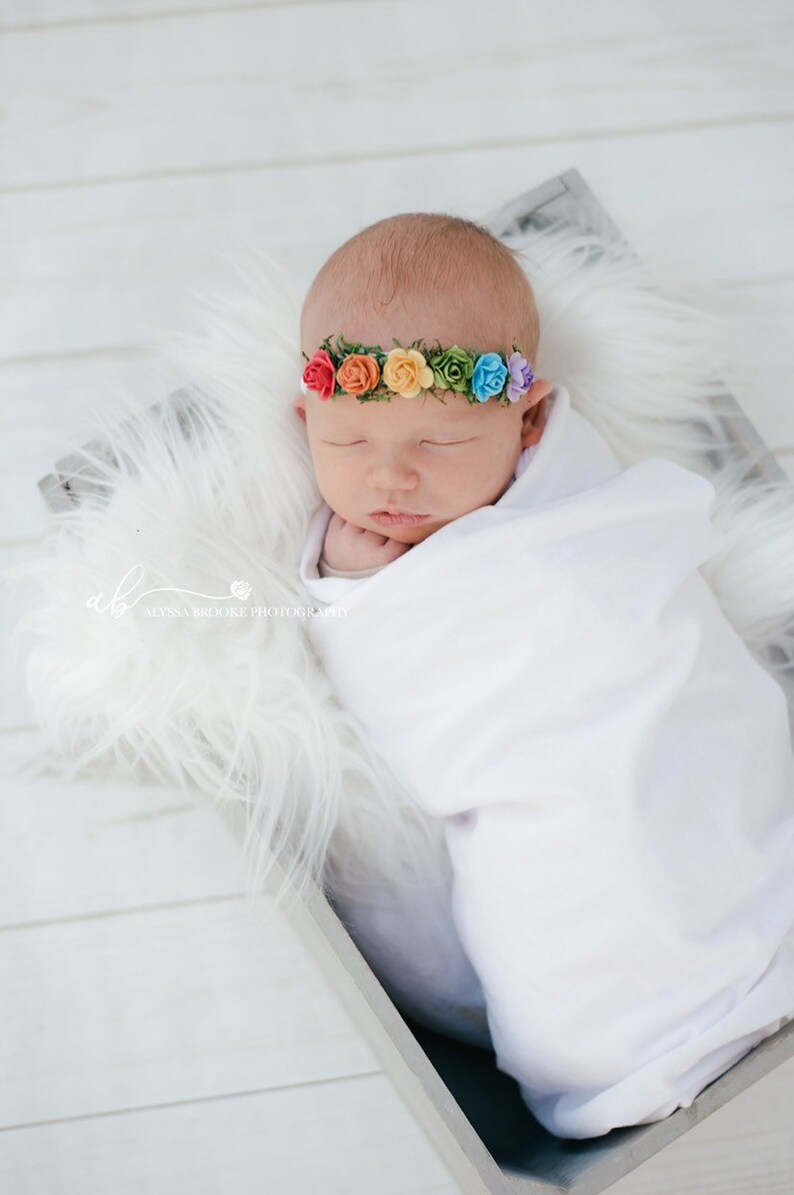 K&ouml;nnte beinhalten: Ein neugeborenes Baby mit einem Regenbogenblumen-Stirnband schl&auml;ft in einem wei&szlig;en, mit Fell ausgekleideten Korb. Das Baby ist in eine wei&szlig;e Decke gewickelt und hat die Augen geschlossen.