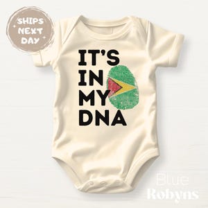 May include: Cream-colored baby bodysuit with the text "IT'S IN MY DNA" in black. A fingerprint design in the colors of the Guyana flag is next to the text. The bodysuit has short sleeves.