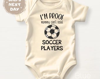 May include: Cream-colored baby bodysuit with the text "I'M PROOF mommy can't resist SOCCER PLAYERS" and a soccer ball graphic. The bodysuit is short-sleeved and has snap closures. The text and graphic are in black.